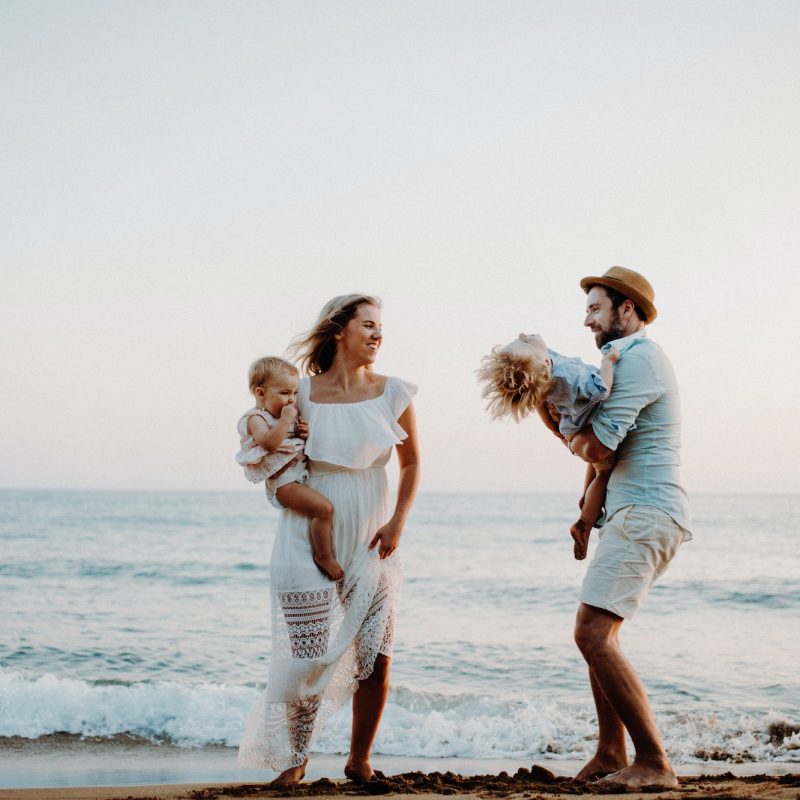 A young family with two toddler children standing on beach on summer holiday.